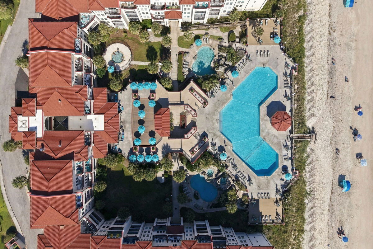 Overhead aerial view of the resort courtyard detailing the central pool, umbrella-lined lounging terraces, secondary water features, and direct beach access paths.