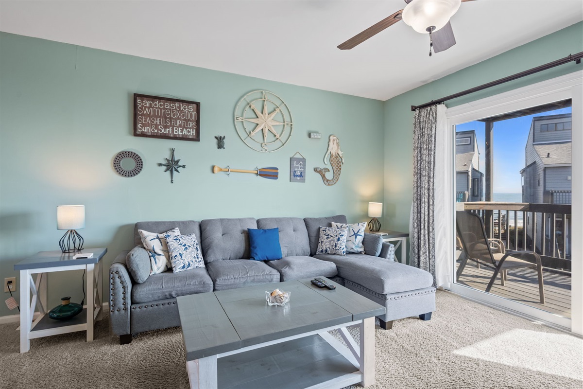 A bright coastal living space designed for unwinding, with a comfy sectional, beach-inspired decor, and sliding doors that open to a balcony view of the water