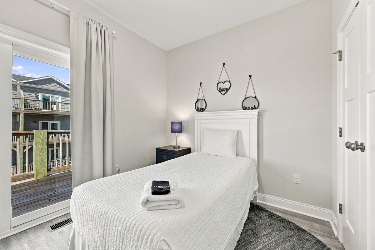 Cozy bedroom featuring a white beadboard headboard, crisp white bedding, decorative wall mirrors, and direct sliding glass door access to a private wooden balcony deck.
