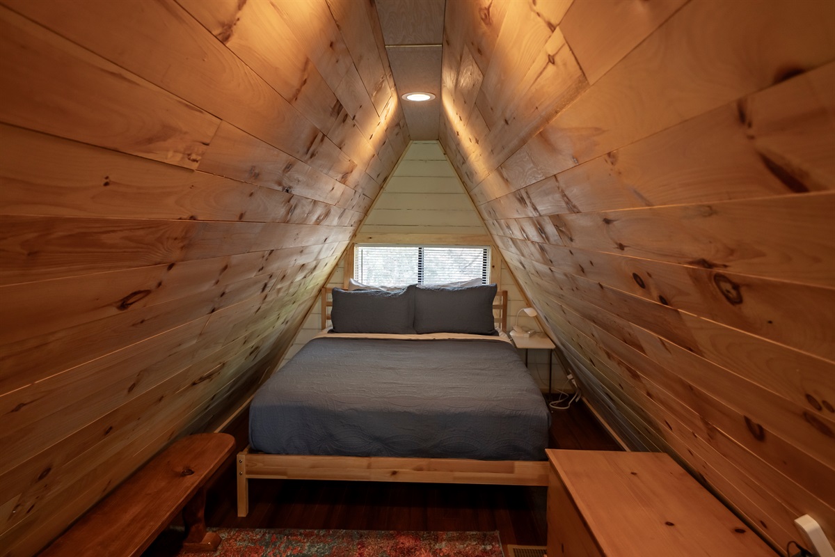 A true A-frame experience—this loft bedroom features a queen bed and a cozy sitting area overlooking the trees through the home’s signature windows.