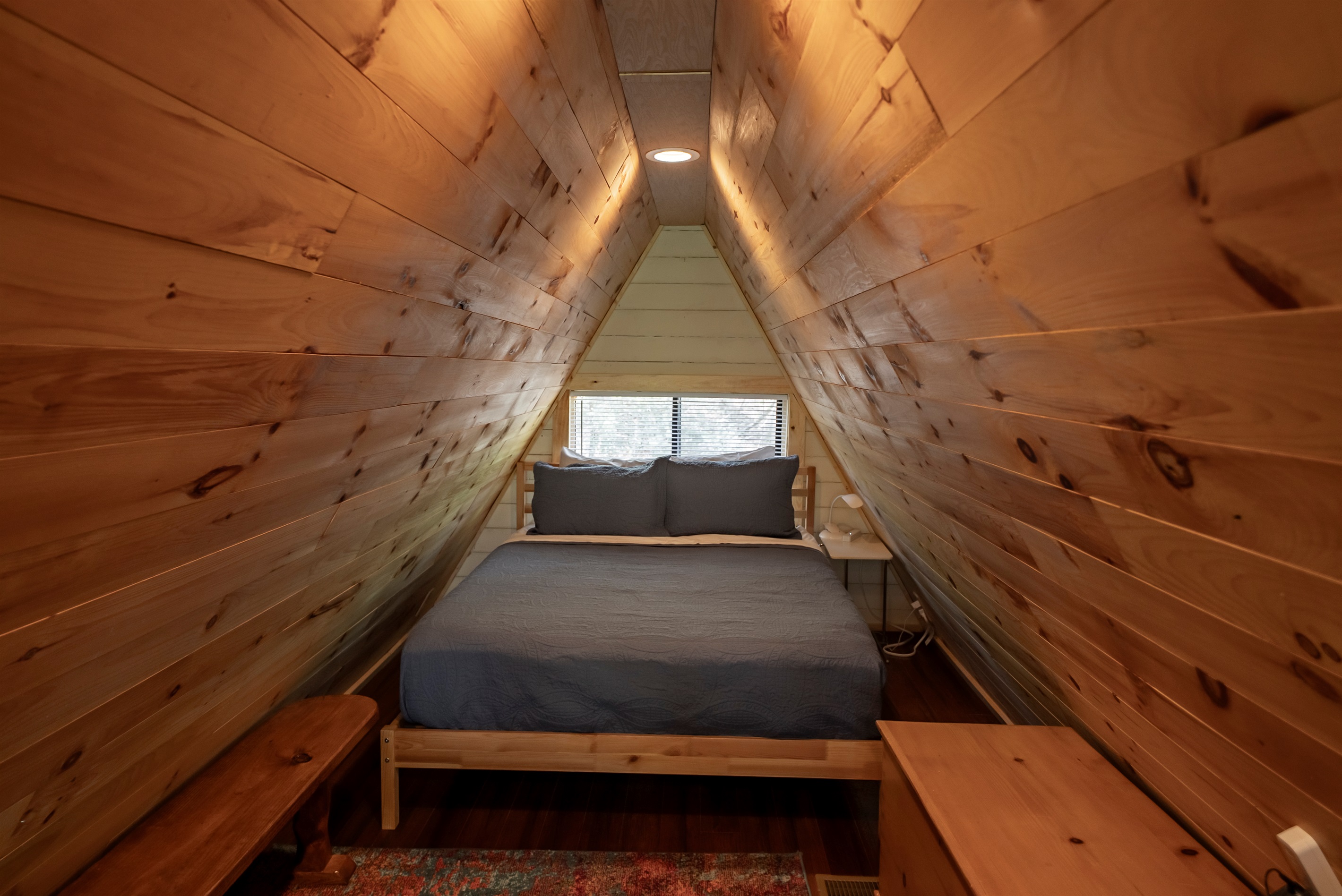 A true A-frame experience—this loft bedroom features a queen bed and a cozy sitting area overlooking the trees through the home’s signature windows.