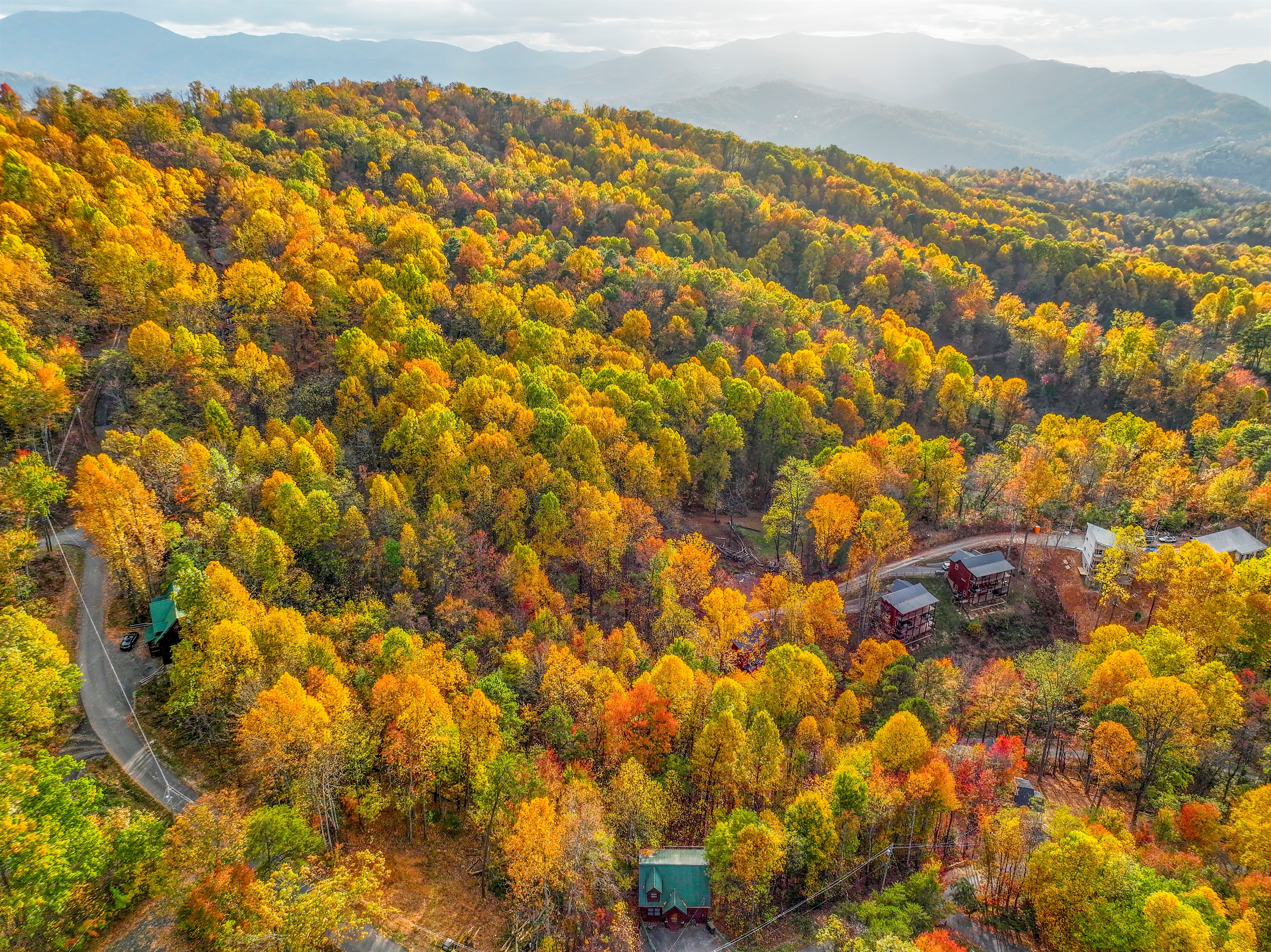 An aerial view of a serene autumn landscape, where vibrant hues of gold, orange, and red blanket the forest. Cozy cabins nestle among the trees, with majestic mountains rising in the distance.