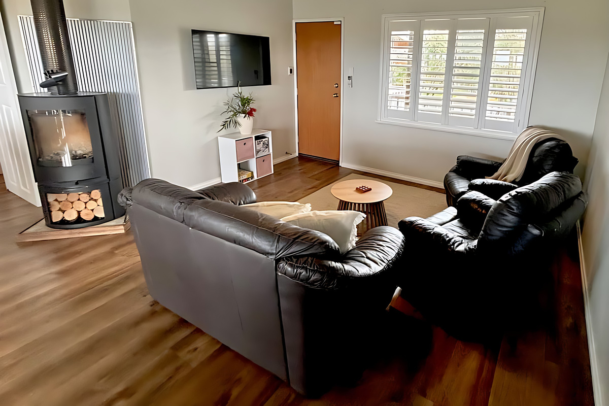 The living room at Lake Views & Coastal Escape has a raked ceiling, a leather lounge and recliners, a wall-mounted smart TV and a freestanding wood fire with a stacked log store beside it.