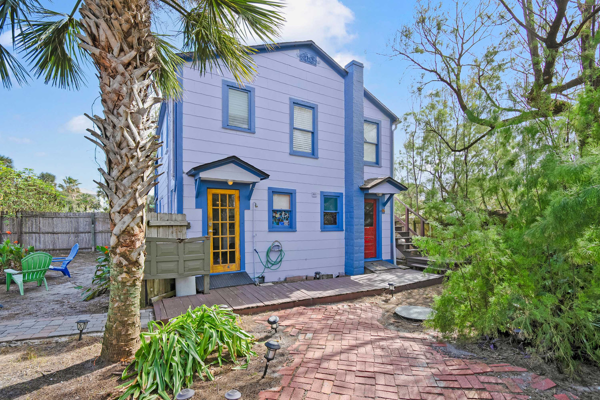 The bungalow seen from the lane, lavender clapboard against the palms, with the exterior stairs up to Unit B over on the right. Photographs never quite catch how green this whole street becomes in high summer.