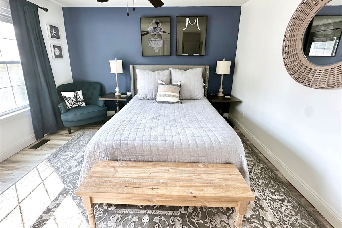This modern bedroom features a blue accent wall and a neutral bed with a wooden end-of-bed bench. The space includes a teal armchair, unique wall art, and a large round mirror, all set over stylish hardwood-style flooring.