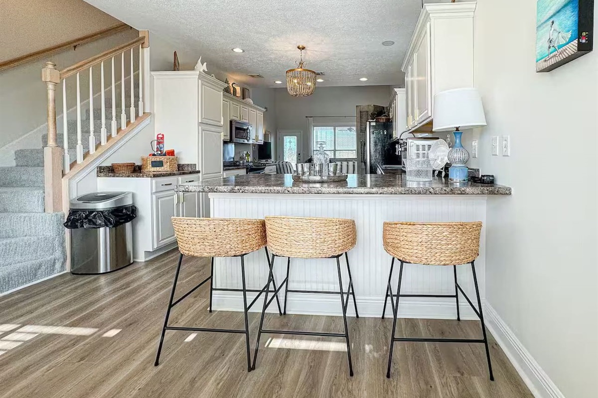Bright, open-concept kitchen with a large island, granite countertops, and three woven bar stools. Features modern white cabinetry, wood-style flooring, and elegant lighting—perfect for cooking and gathering.