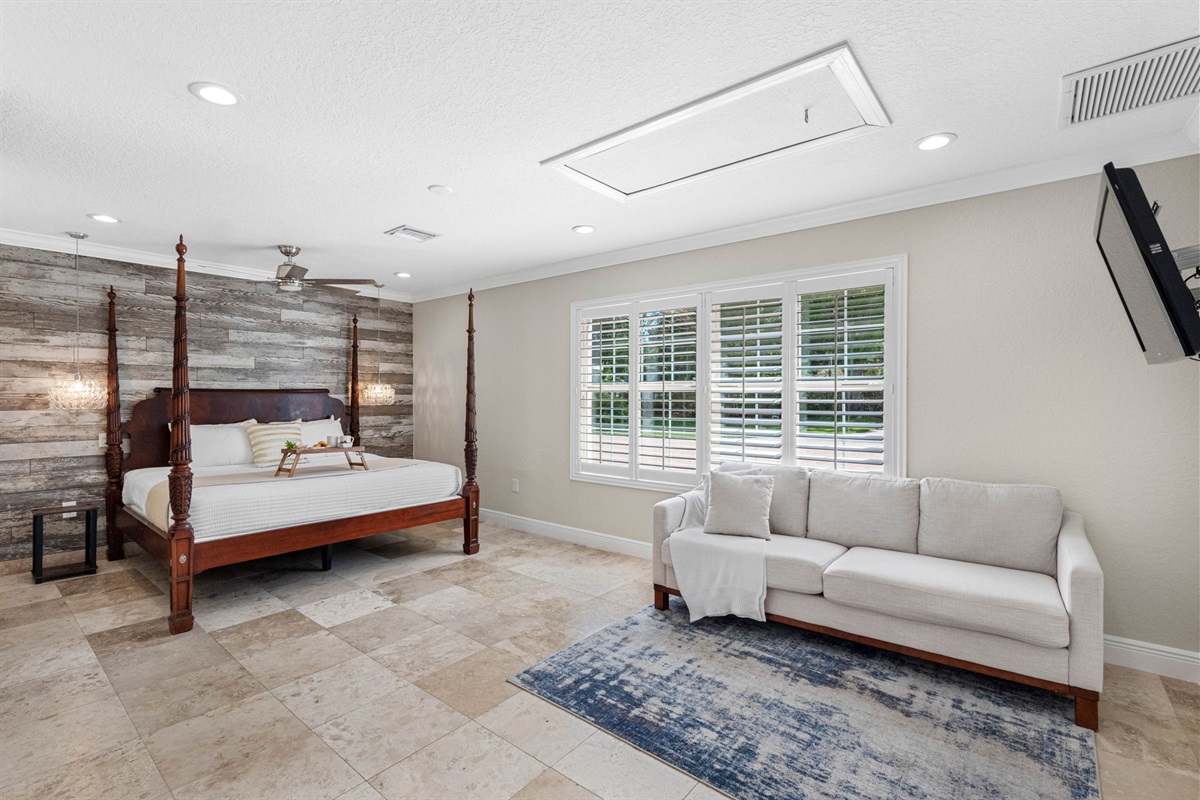 This inviting master bedroom features a grand four-poster bed, a rustic wood accent wall, and ample natural light streaming through plantation shutters.