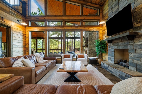 This stunning cabin living room features floor-to-ceiling windows, a stone fireplace, and leather sofas for the perfect cozy retreat. Enjoy the modern rustic design and serene nature views in this luxury cabin rental.