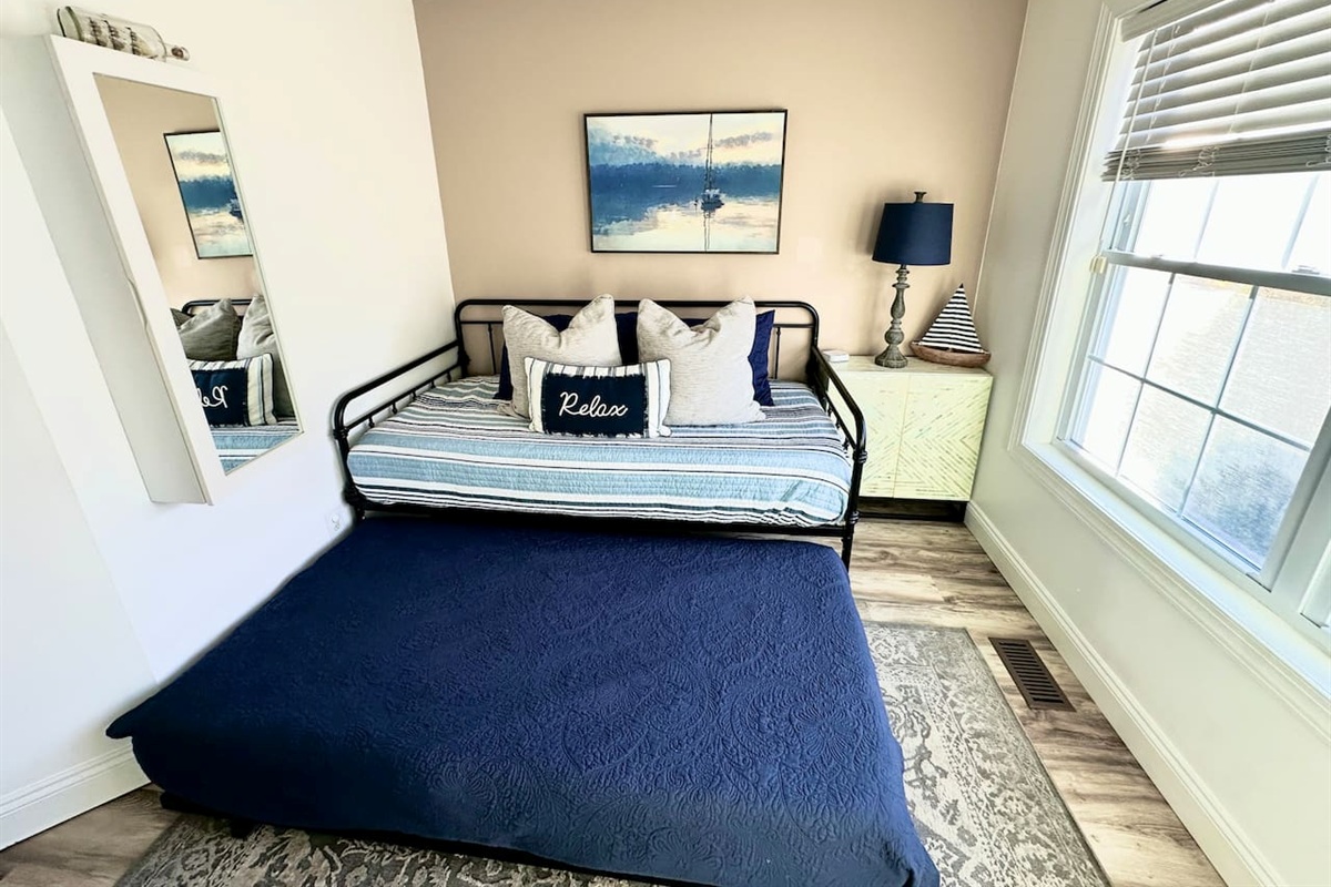 This functional room features a black metal daybed with a pull-out trundle bed dressed in a navy textured coverlet. The space includes nautical wall art, a light-green cabinet with a lamp, and a large window with blinds.
