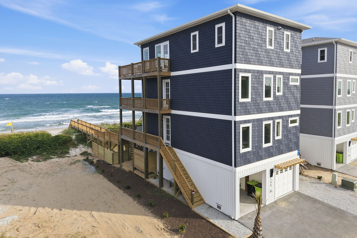 Elevated exterior view showing the oceanfront home&rsquo;s side elevation, connecting walkways, neighboring properties, and direct beach proximity with coastal dune landscape