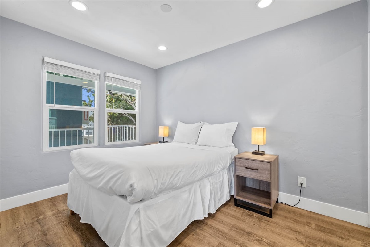 Inviting bedroom with a comfortable bed, flanked by two elegant lampshades, offering soft lighting. A large window brightens the space, and the aesthetic flooring adds warmth, creating a cozy and restful retreat.