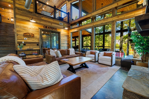With high ceilings, open floor plan, and natural lighting, this luxury cabin living space offers warmth and comfort. Plush leather seating and a fireplace create a modern rustic home, perfect for a vacation retreat in nature.