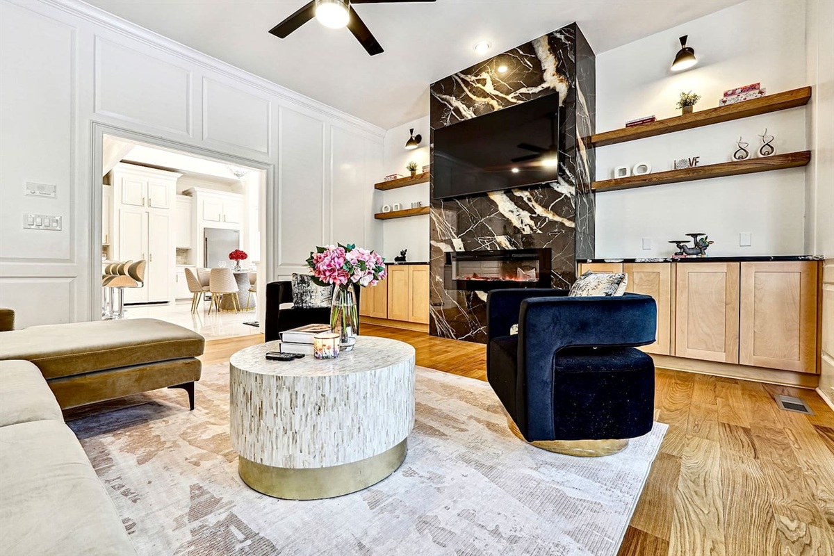Situated next to the kitchen, this main-floor living room offers a warm, upscale ambiance with a brown L-shaped sectional, two black accent chairs, gold and black design accents, a smart TV, and a fireplace, perfect for entertaining or unwinding.