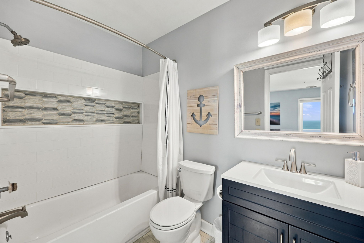 En-suite guest bathroom featuring a navy blue vanity with white countertop, brushed nickel hardware, rustic framed mirror with overhead vanity lighting, anchor wall accent, and a tiled shower-tub combination with a textured mosaic inlay niche.   