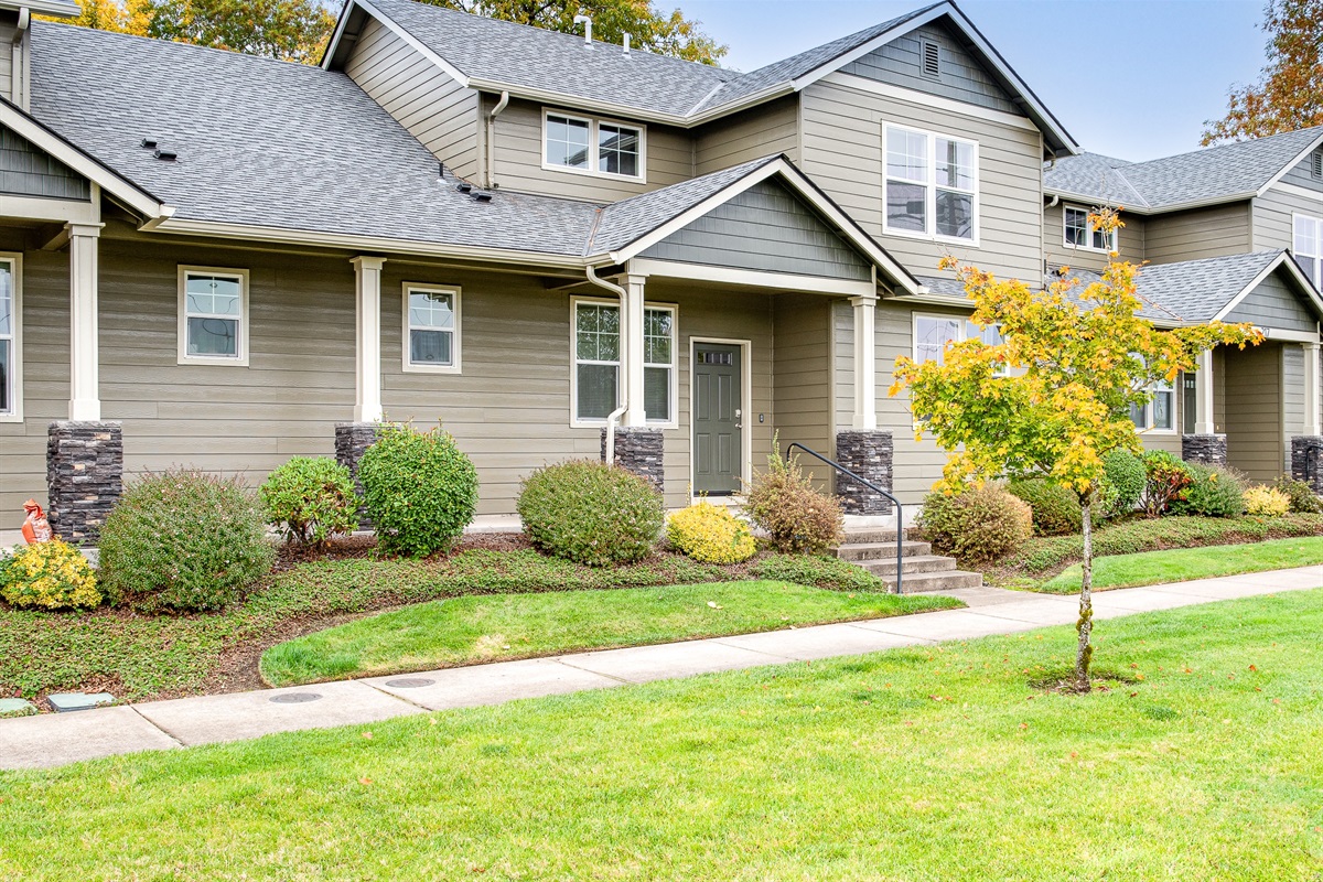 A modern two-story townhouse with a gray exterior, featuring white trim, a front porch with stone columns, and well-maintained landscaping including bushes and a small tree.