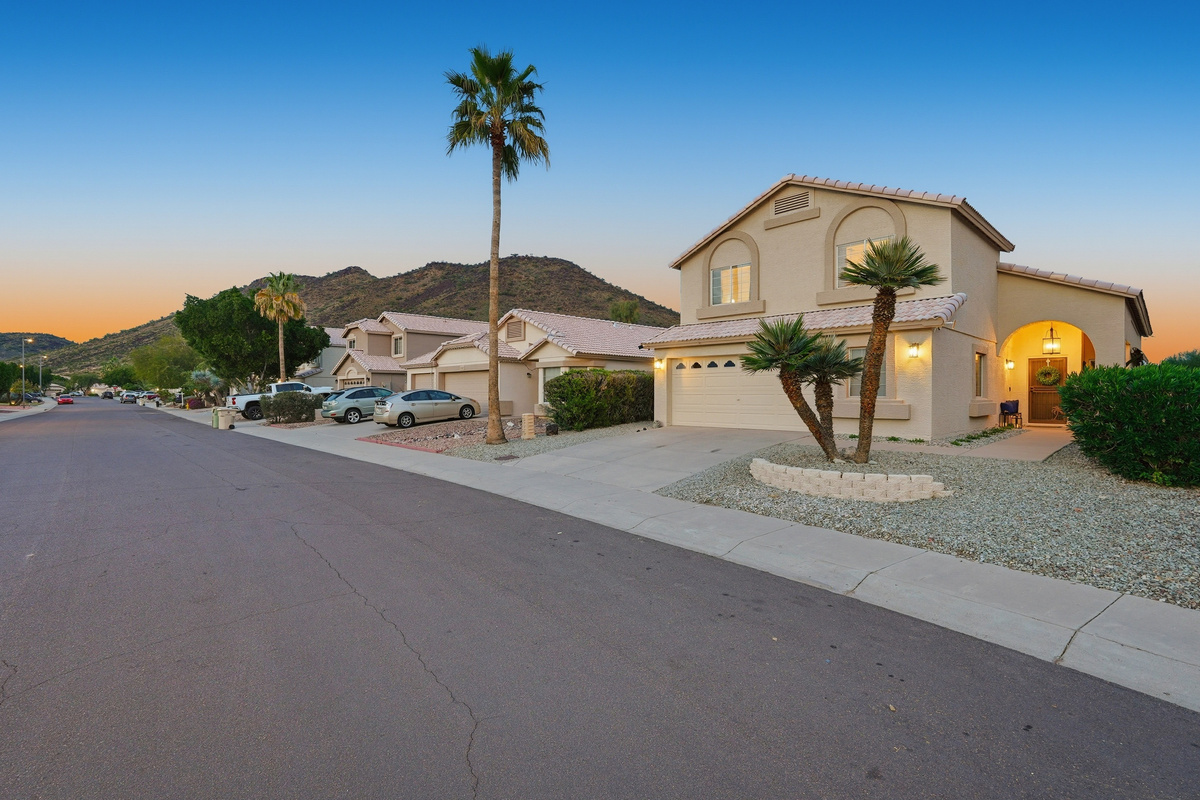 Front exterior at dusk with driveway parking, desert landscaping, palm trees, and mountain views nearby.