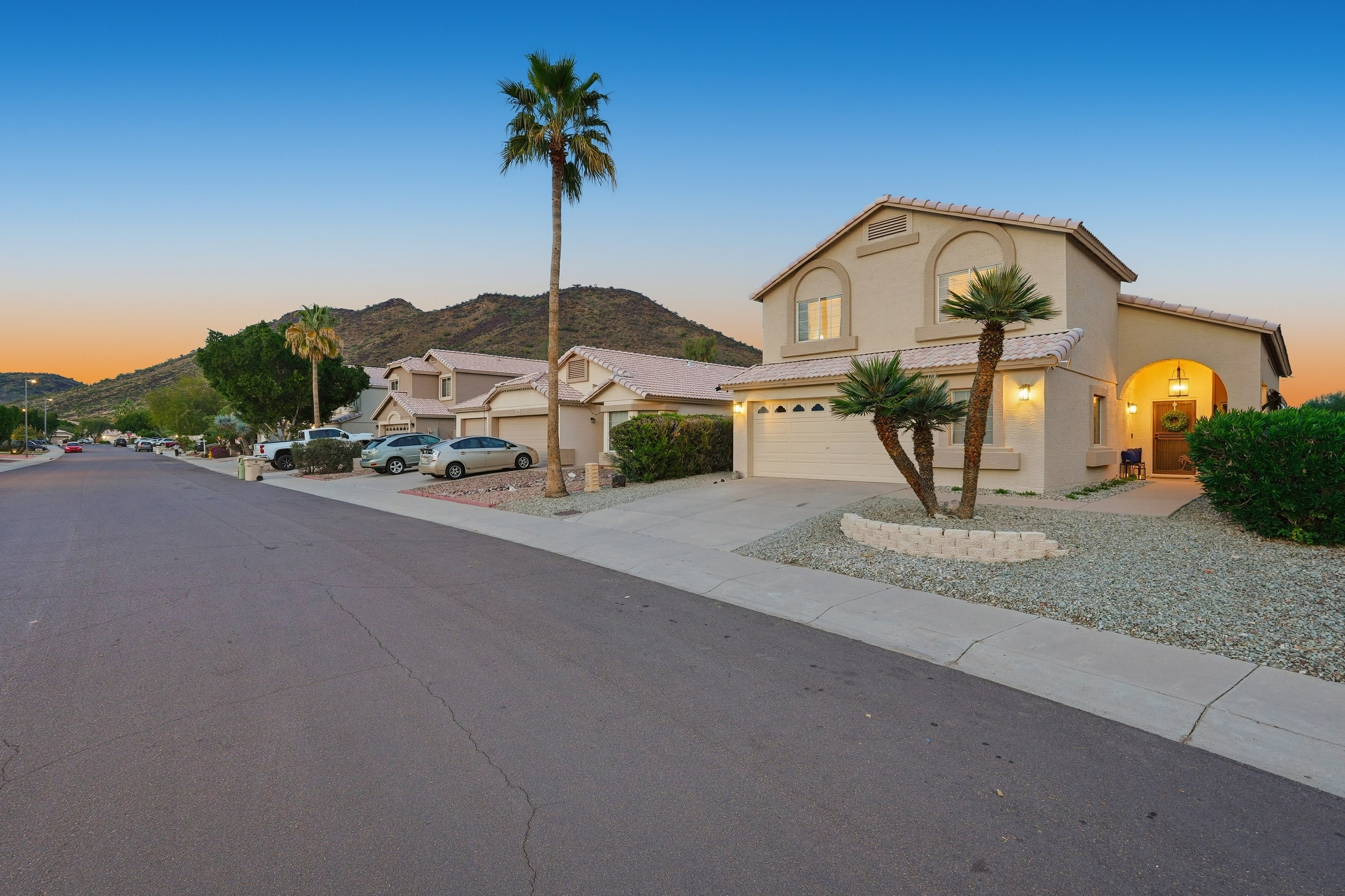 Front exterior at dusk with driveway parking, desert landscaping, palm trees, and mountain views nearby.