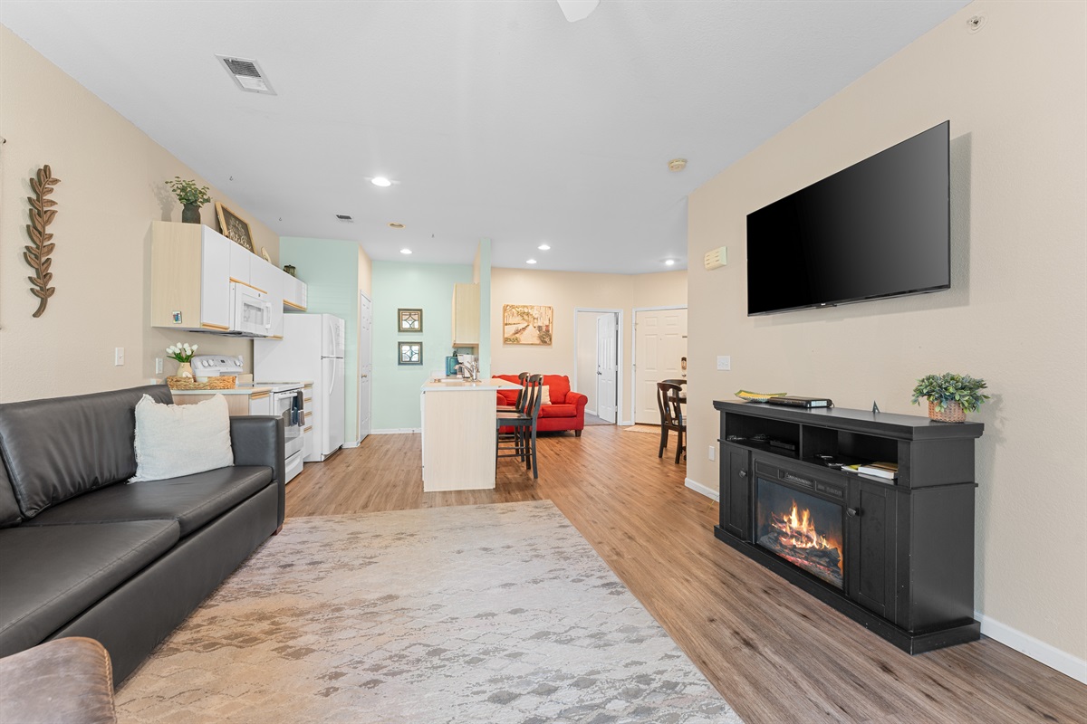 Reverse living-room angle showing the fireplace, TV, and connection to the kitchen and entry.