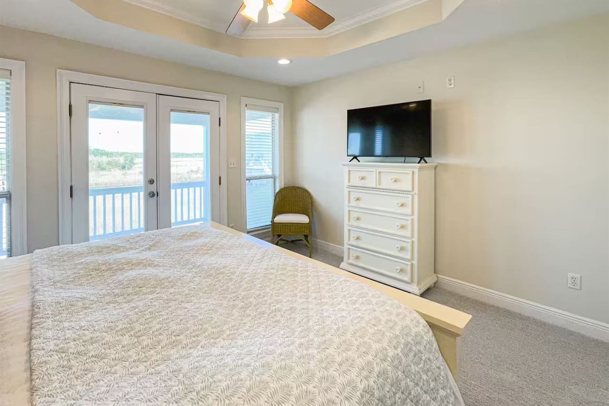 Airy bedroom with a comfortable bed, flat-screen TV, and private balcony access. Features abundant natural light, ceiling fan, and a calm, coastal feel.