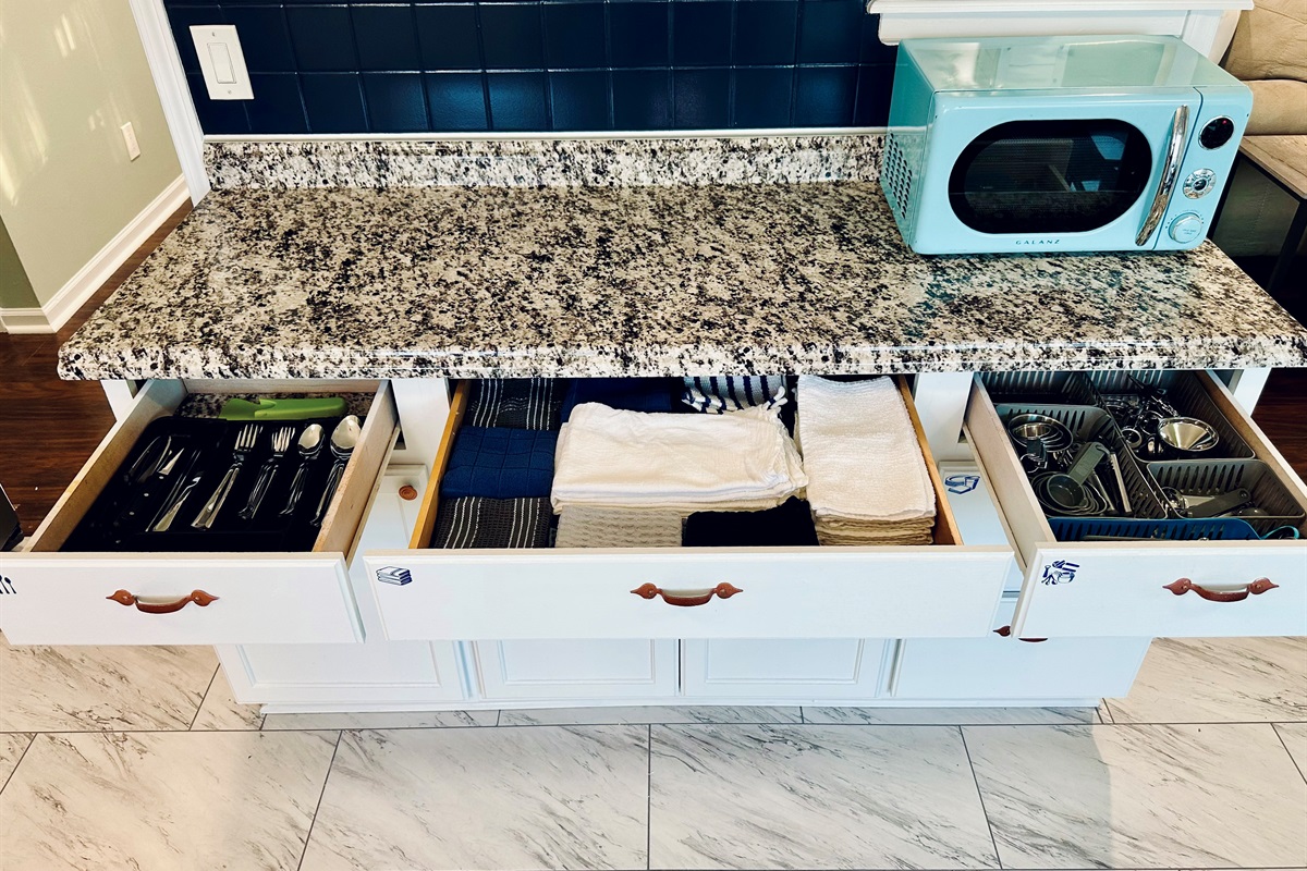 Kitchen drawers organized with a full set of silverware and a variety of kitchen towels, essential cooking tools, including measuring cups, measuring spoons, can opener, vegetable peeler, grater, and other utensils, neatly arranged and easy to access.