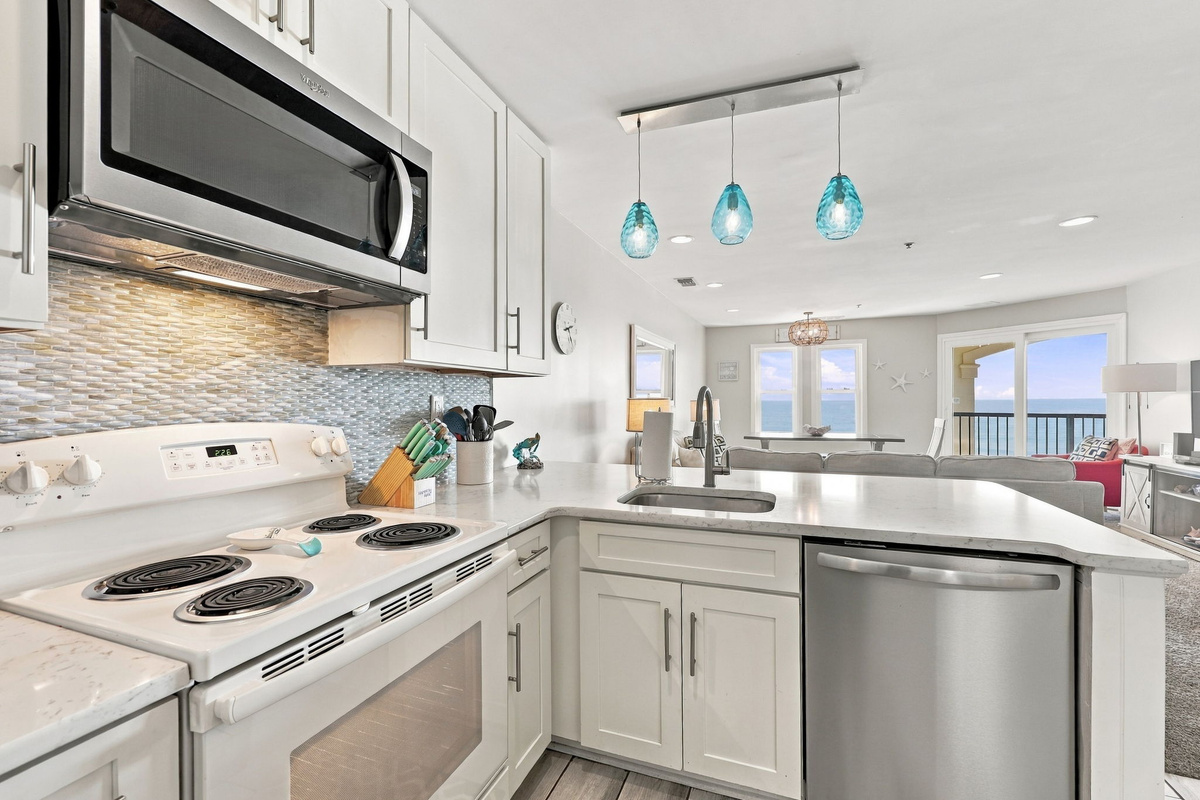 Close-up perspective of the modern coastal kitchen highlighting the polished white quartz countertops, sleek stainless steel dishwasher, undermount sink, built-in microwave, white shaker cabinetry, and glowing aqua teardrop pendant lights framed against sw