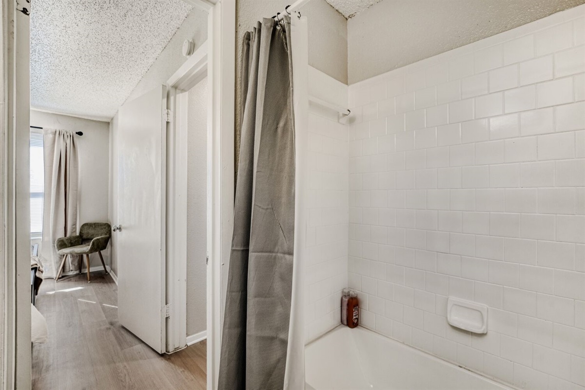 Bright bathroom with white tile walls, a shower-tub combo, and a view into a cozy bedroom.