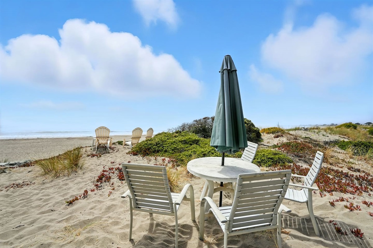 Outdoor dining set on sandy beach with umbrella shade.

