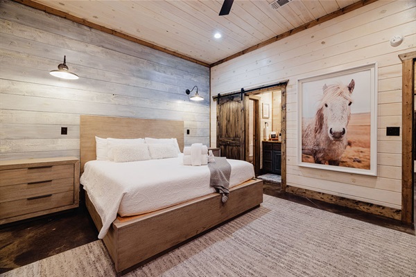 Charming farmhouse-style bedroom with a wooden king-size bed, rustic décor, a barn door entrance to an en-suite bath, and warm lighting. Ideal for a luxury cabin experience.