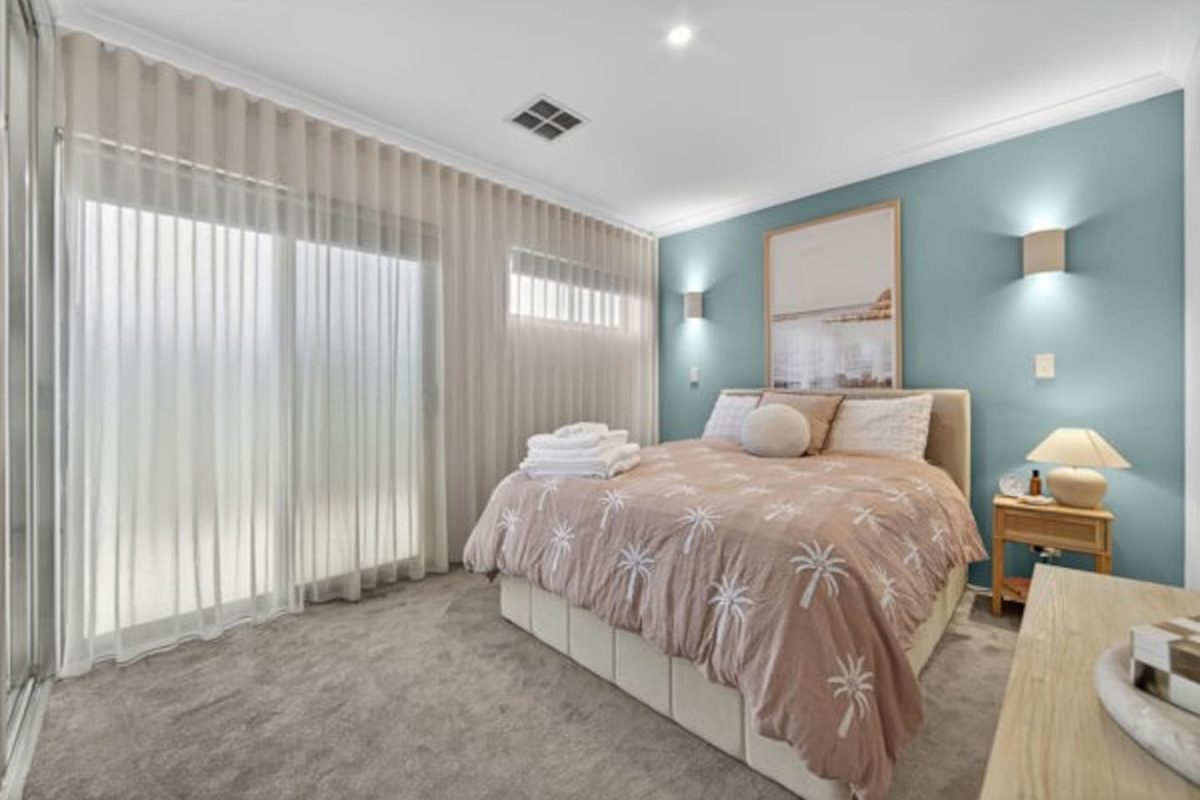 Light, airy bedroom with plush carpet, floor-to-ceiling sheers, ducted air conditioning and a built-in robe, with fresh towels ready on the bed.