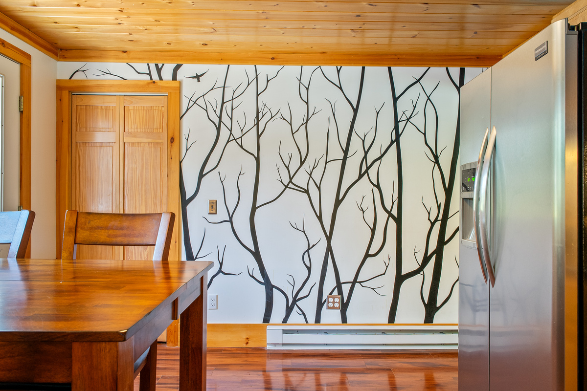 Look across the dining area to reveal the stainless steel side-by-side refrigerator equipped with an exterior ice and water dispenser. The shot highlights the custom tree accent wall, hardwood flooring, and bifold doors to the kitchen pantry.