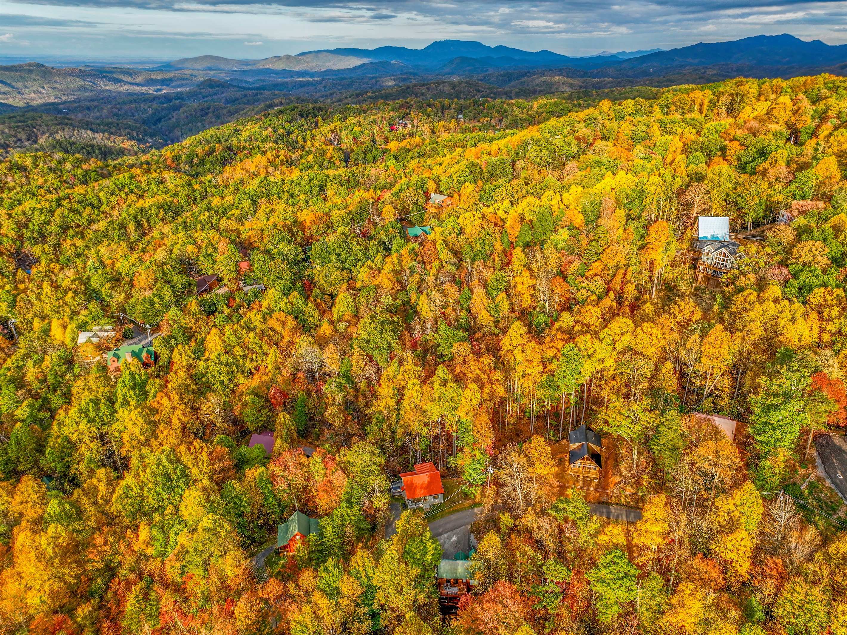 An aerial view of a serene autumn landscape, where vibrant hues of gold, orange, and red blanket the forest. Cozy cabins nestle among the trees, with majestic mountains rising in the distance.