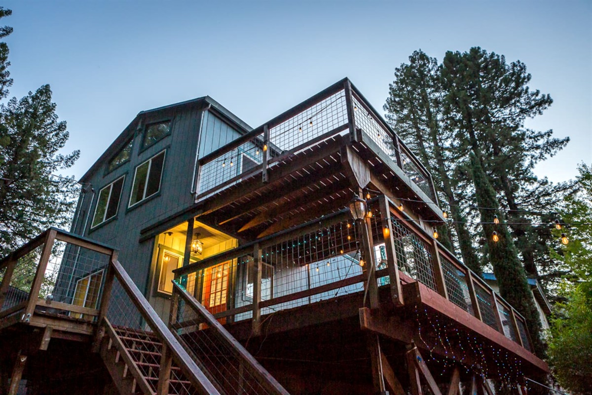 - Two story home with wraparound decks adorned with warm glowing string lights  
- Elevated design provides scenic views of surrounding towering pine trees  
- Multiple outdoor seating areas are perfect for relaxing or hosting guests outdoors  
