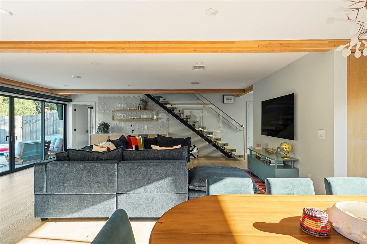 A spacious open-concept living area featuring exposed wooden beams, a large sectional sofa, a modern staircase with a glass railing, and sliding doors that open to an outdoor space.