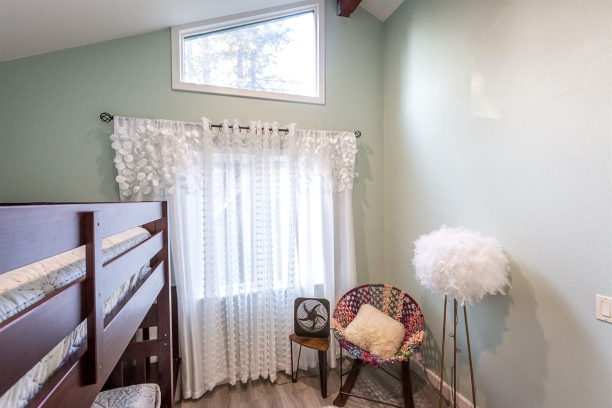 - Cozy bedroom corner with wooden bunk beds and colorful accent chair  
- Soft natural light through sheer curtains creates a gentle atmosphere  
- High ceiling with transom window adds openness to the intimate space  