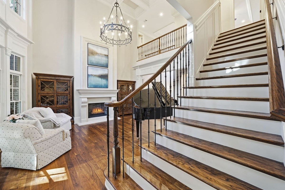 Beautiful entry sitting room with a grand piano, warm fireplace, tall windows, and two comfortable chairs. A bright, serene spot to unwind or enjoy quiet moments.