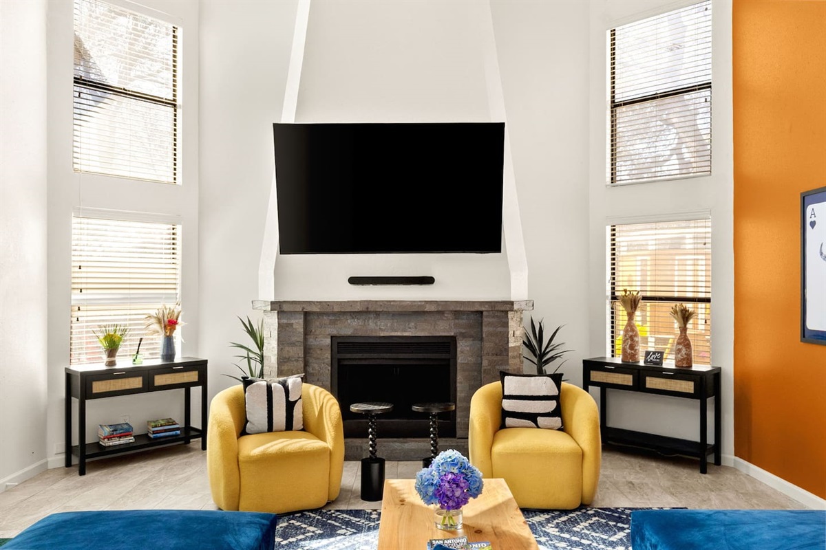 Bright and stylish living room with cozy yellow chairs, a sleek fireplace, and oversized TV framed by tall windows. Perfect for movie nights, morning coffee, or relaxing after exploring San Antonio.
