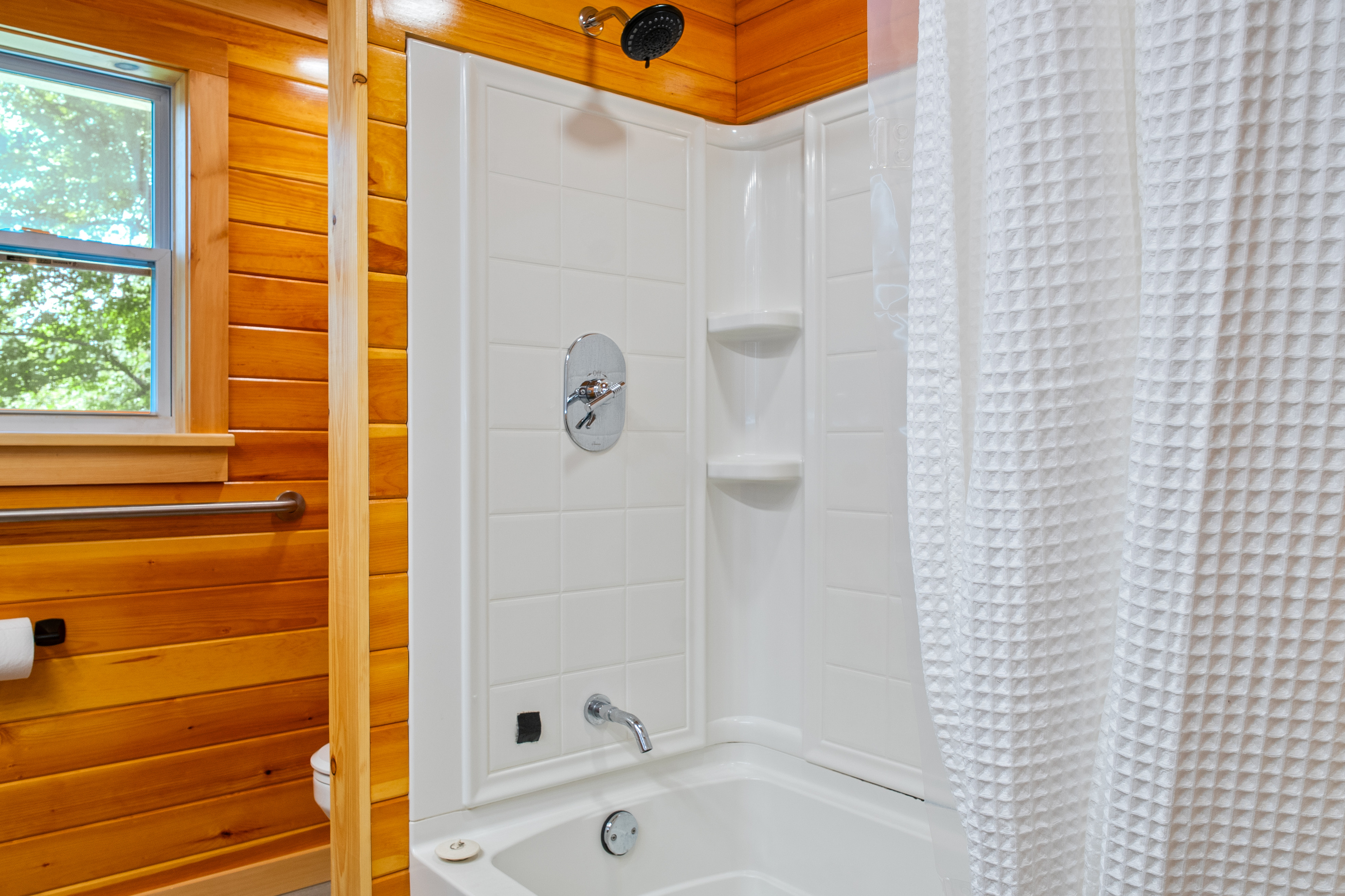 Enjoy a clean, sparkling bathtub and shower combo equipped with built-in corner shelving for your toiletries. This bright station features a modern adjustable showerhead, high-walled white enclosure inserts, and an adjacent window view.