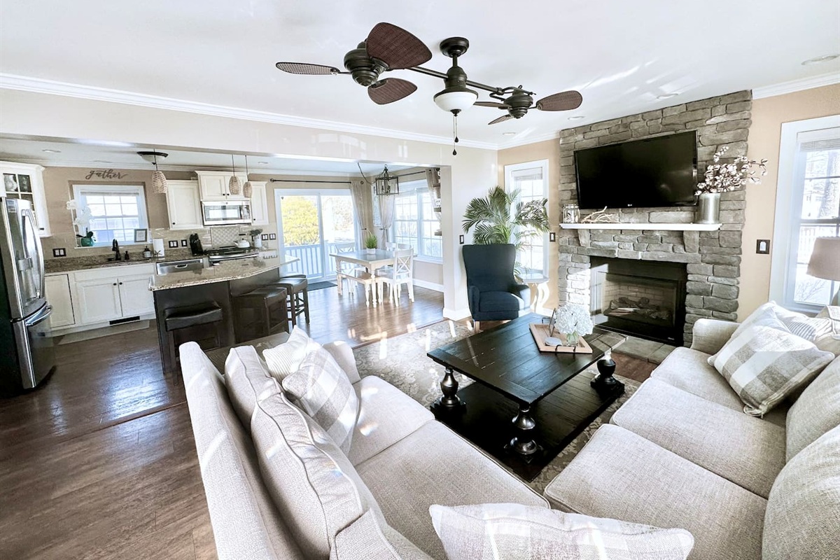 This wide view showcases the seamless flow between the cozy living room, modern kitchen, and dining area. The open-concept design is anchored by a stone fireplace and a unique ceiling fan, creating a spacious and unified gathering spot.