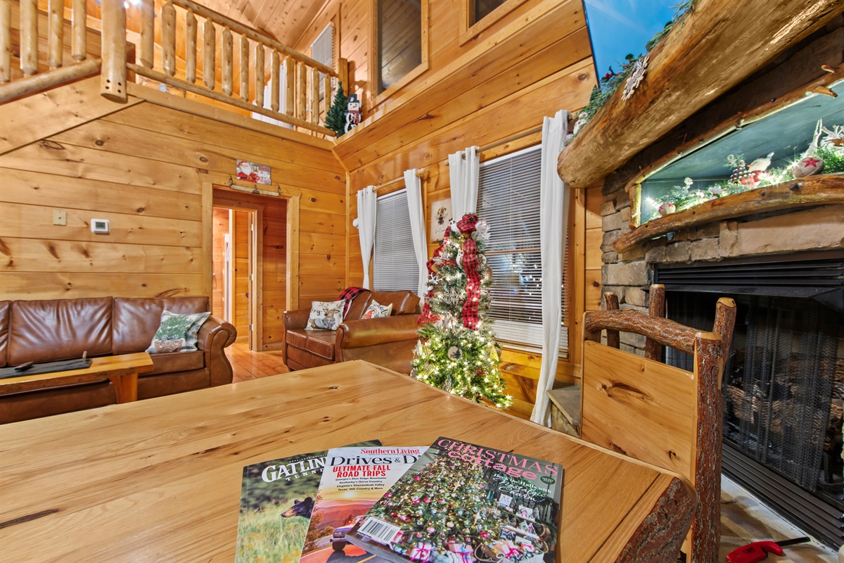 The cozy living room features warm wood paneling, a stone fireplace adorned with rustic decorations, and a leather couch with pillows, creating a welcoming cabin retreat.