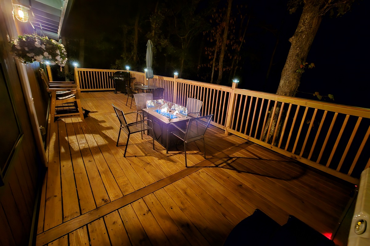 Deck at night featuring fire table, seating, grill, and wooded surroundings.