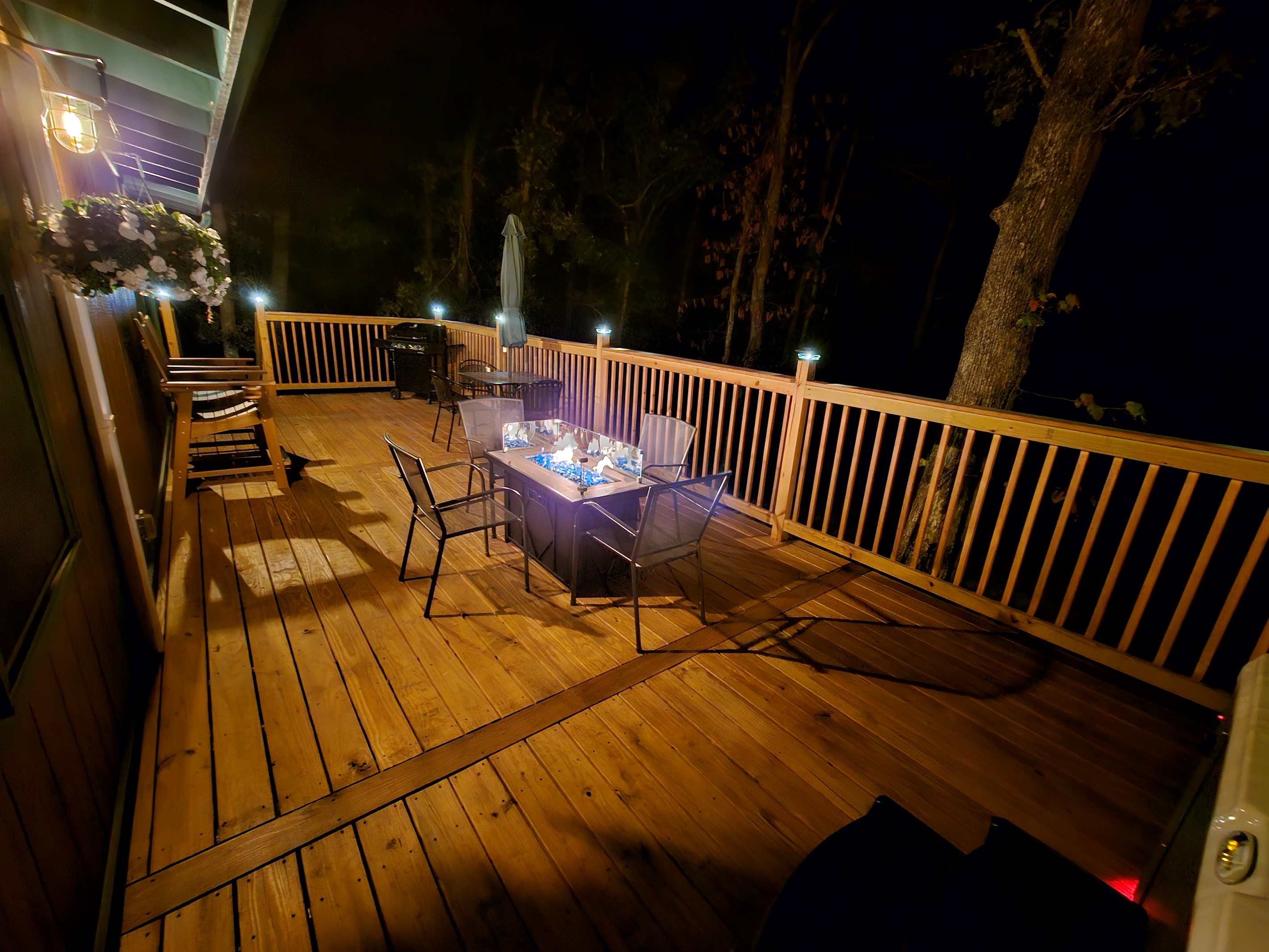 Deck at night featuring fire table, seating, grill, and wooded surroundings.