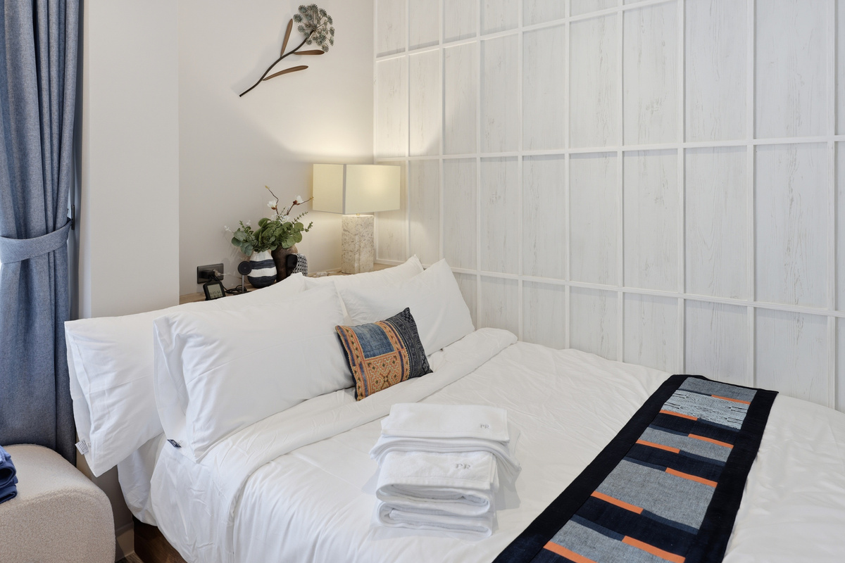 Bedroom with white paneled headboard, white bedding, nightstand with lamp and plant decor, and blue curtains.