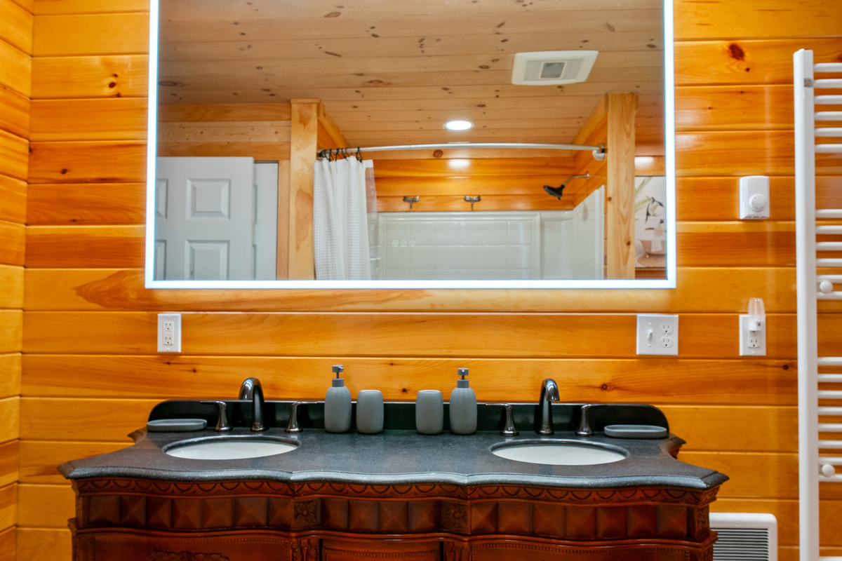 Prepare for the day at the full bathroom's dual vanity featuring two sinks and a modern backlit mirror. Natural pine wall panels surround the space, which includes a shower and bathtub combination visible on the single-story main floor.