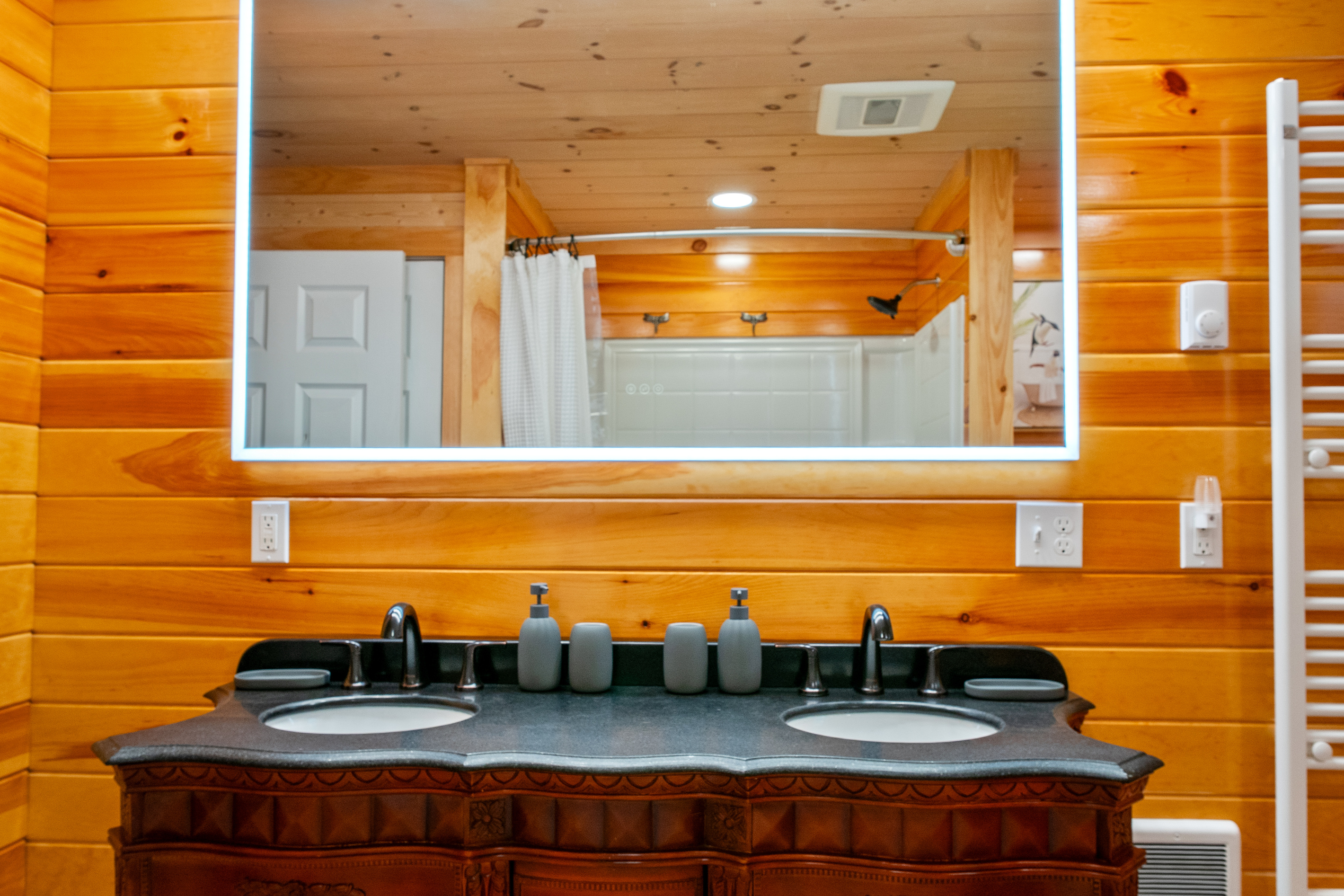 Prepare for the day at the full bathroom's dual vanity featuring two sinks and a modern backlit mirror. Natural pine wall panels surround the space, which includes a shower and bathtub combination visible on the single-story main floor.