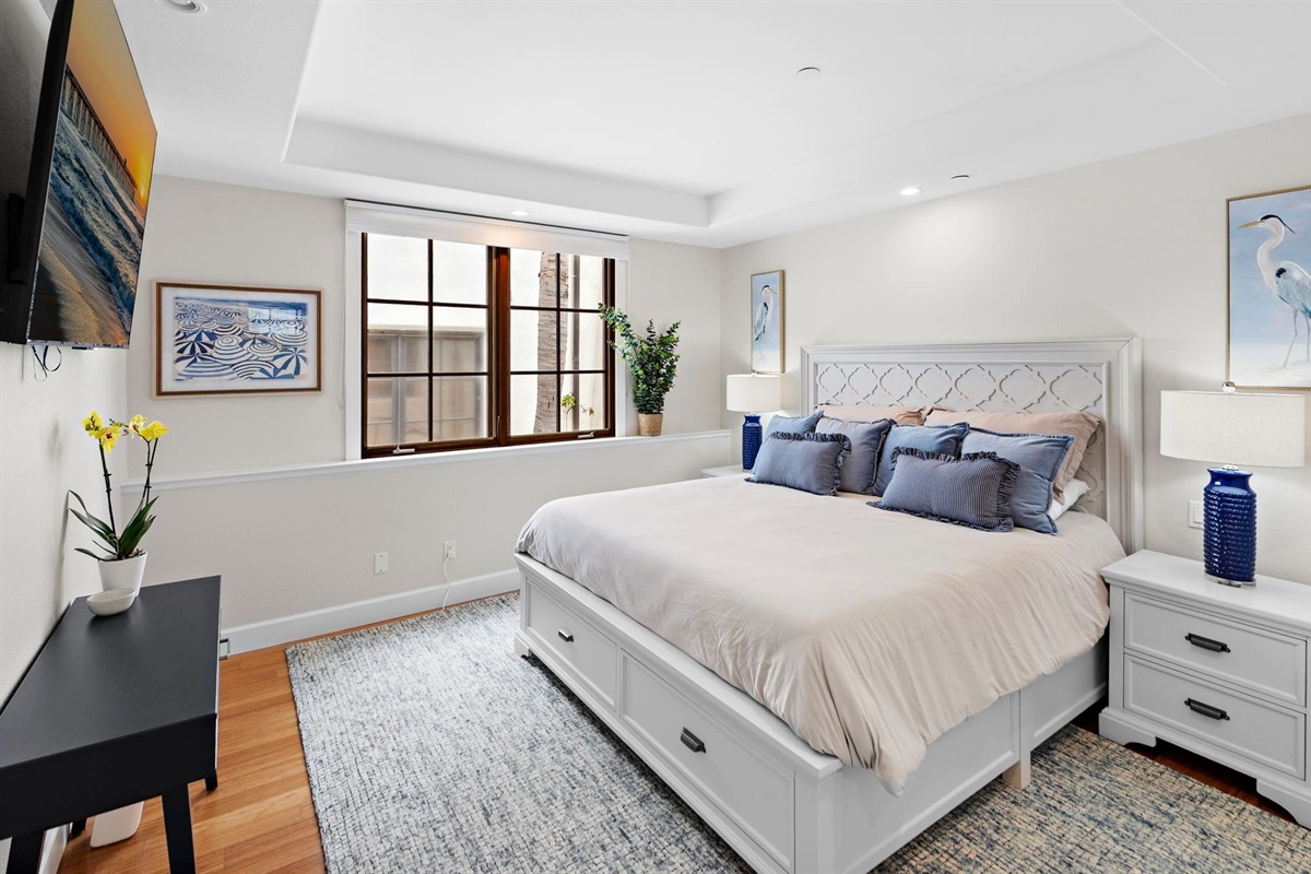 Enjoy a cozy primary bedroom with a plush king bed with premium linens, calming coastal décor, abundant natural light, and a large smart TV for winding down after a day at the beach