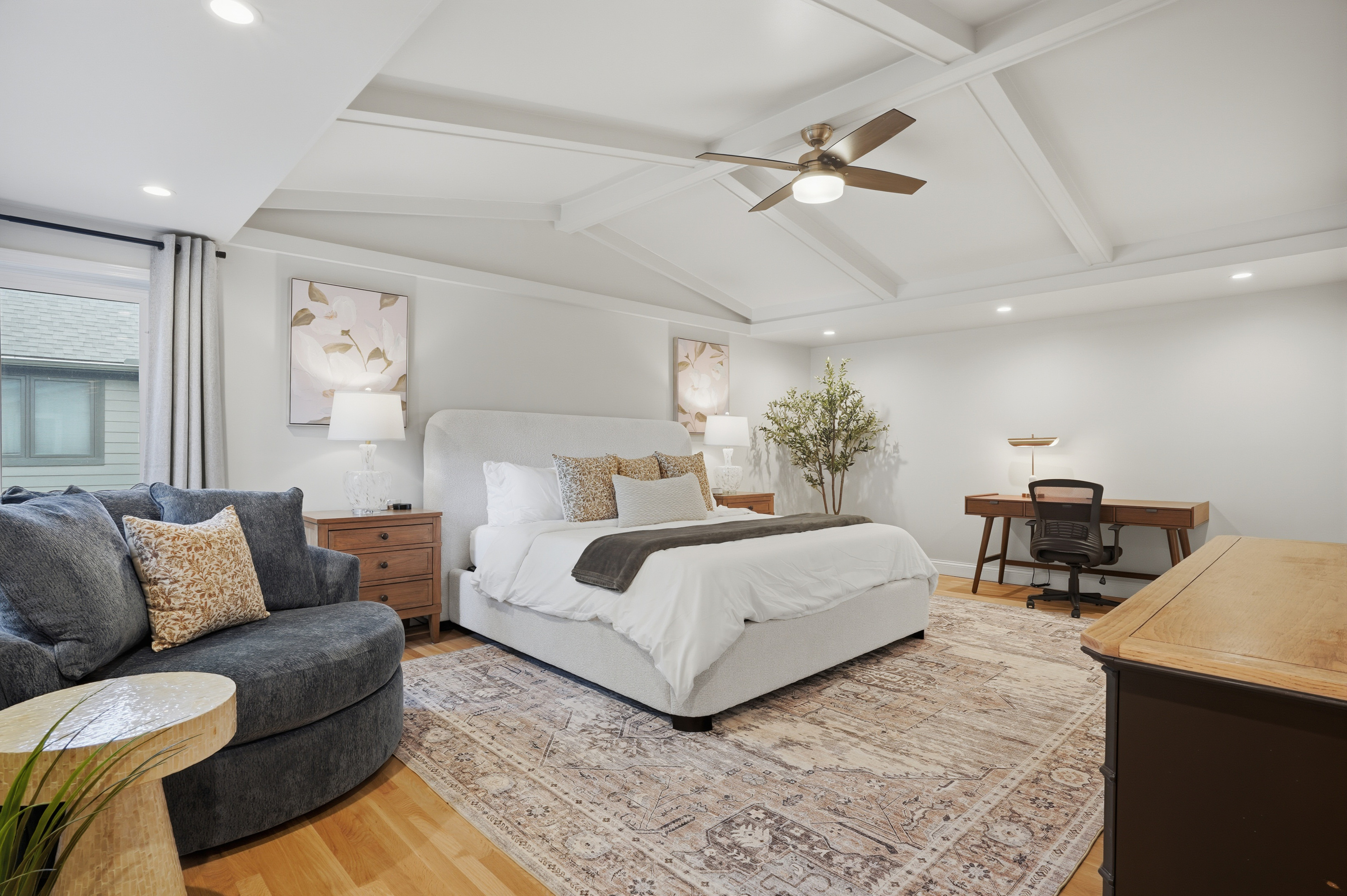 Relax in this spacious primary suite featuring a plush bed, vaulted ceiling with modern fan, natural light, and a comfortable work and seating area designed for both rest and productivity.
