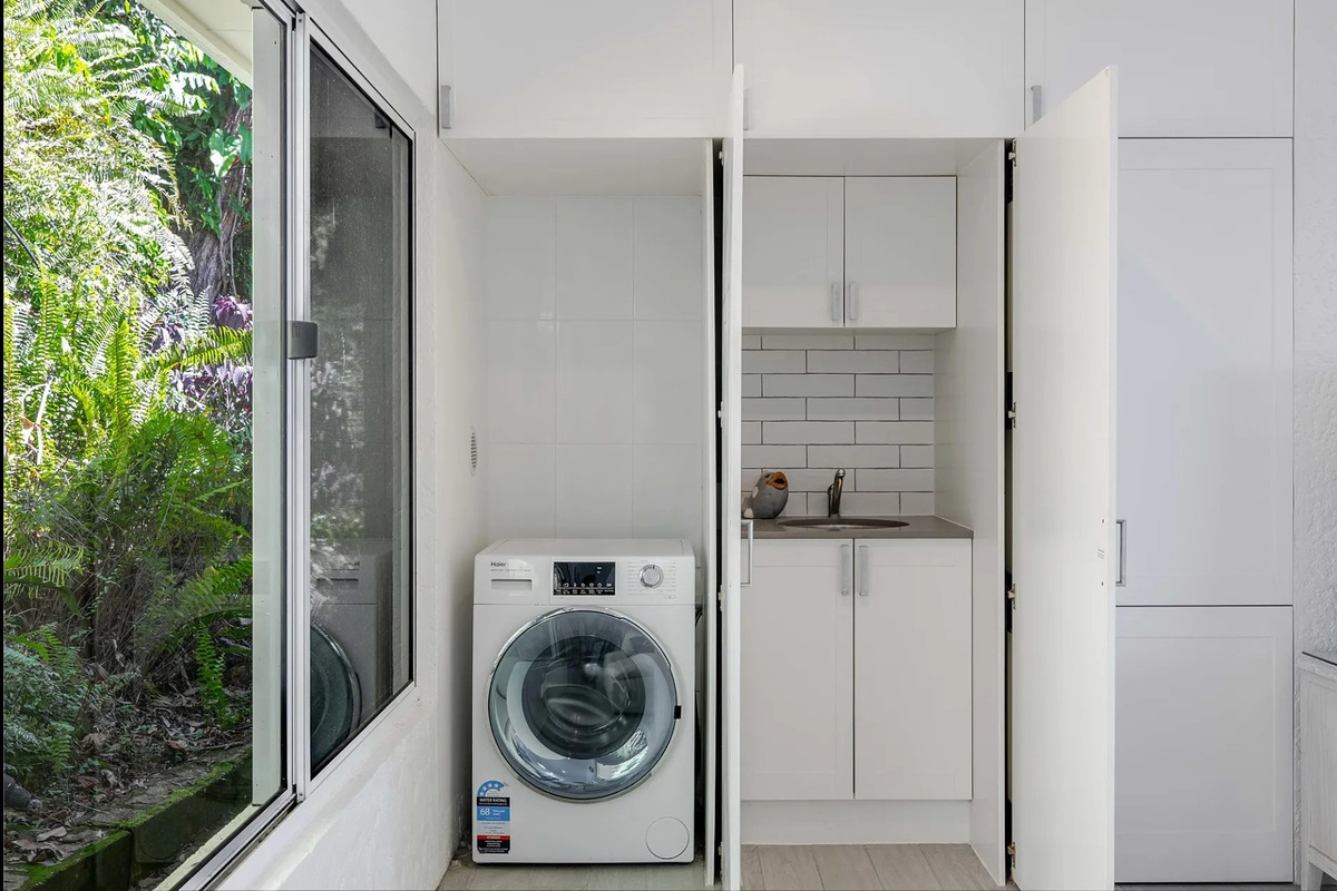 The laundry area includes a front-loader washing machine, storage cupboards and a compact sink tucked neatly beside the garden outlook.