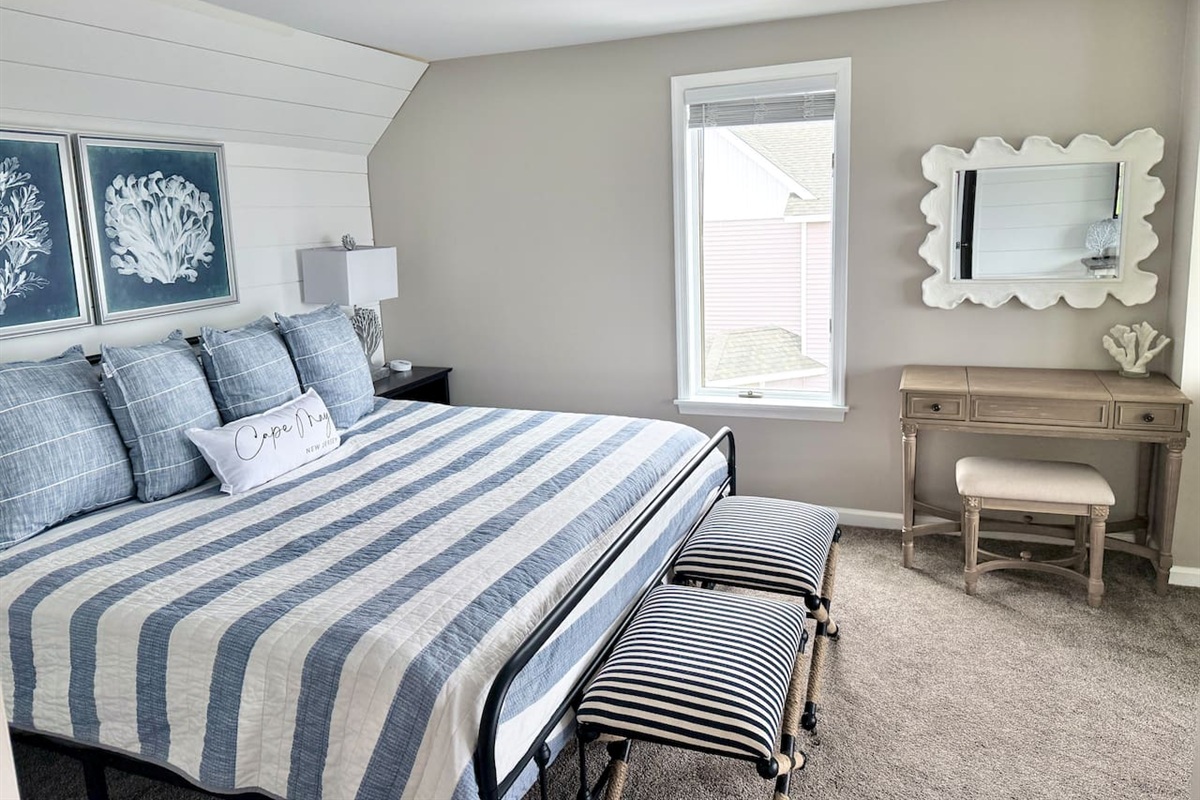 This bedroom features a cozy bed with blue and white striped bedding. The space is accented by a vanity with a unique, textured mirror and a matching stool, completing the tranquil, coastal-inspired design.