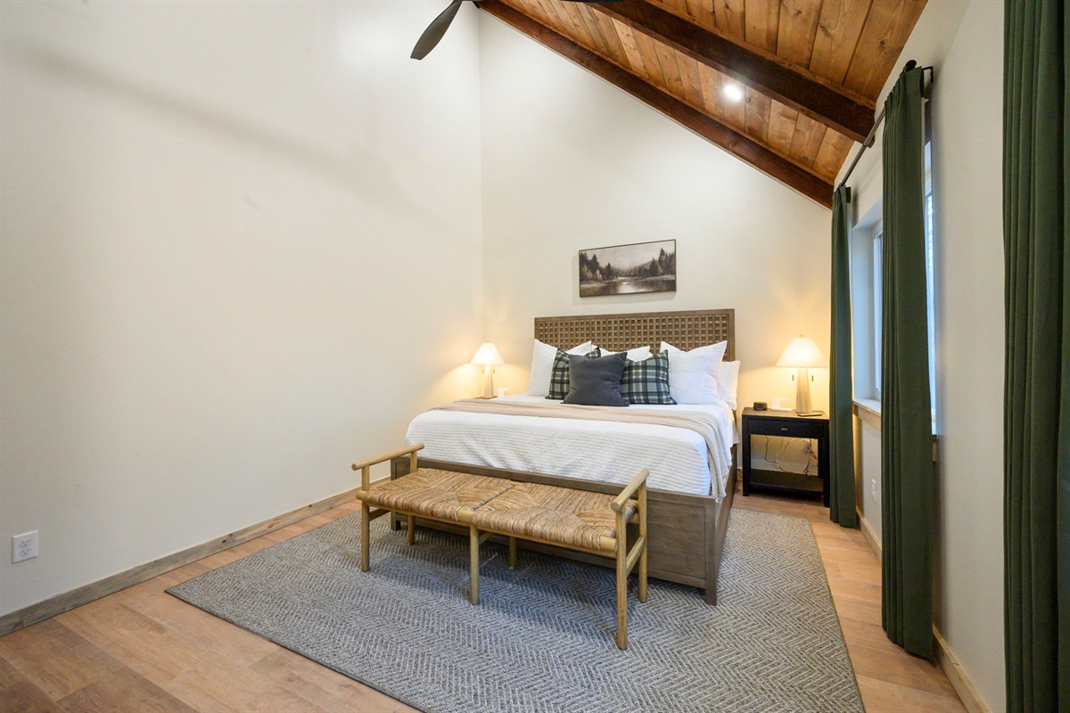 Spacious king bedroom with vaulted ceilings, warm wood beams, and peaceful forest views.