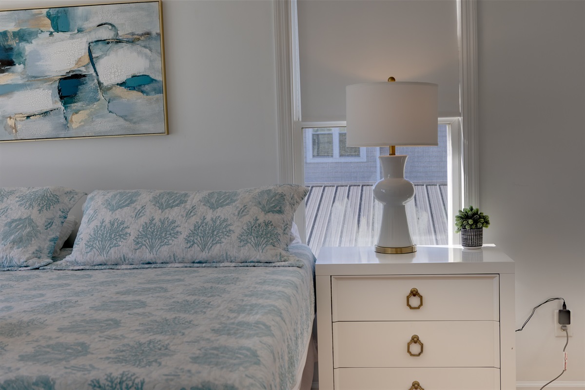 Thoughtfully styled with coastal touches, this upstairs master offers a cozy nightstand with lamp, soft natural light, and fresh, comfortable bedding